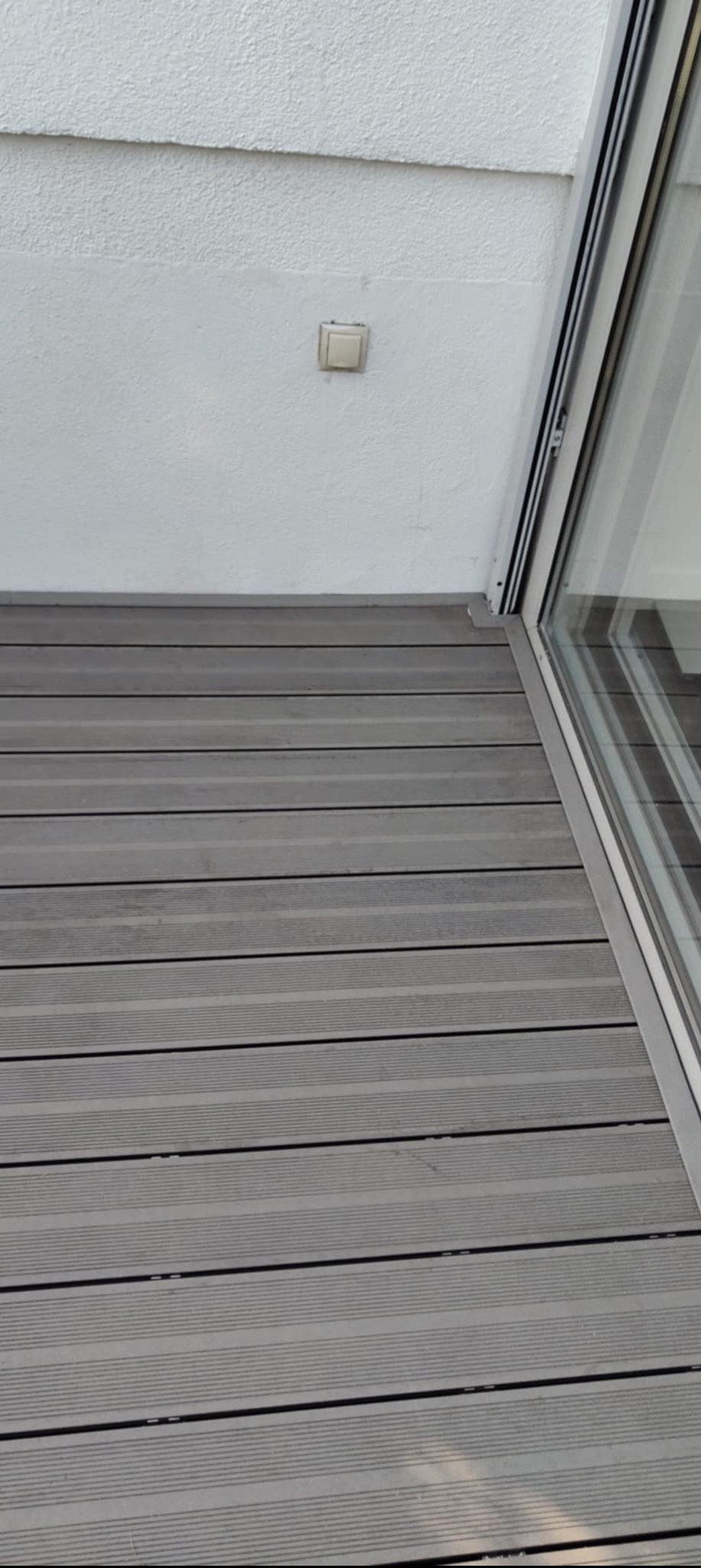 Szare deski tarasowe kompozytowe ułożone na balkonie, widok z góry, fragment okna balkonowego, biała ściana z włącznikiem. Detal wykończenia.