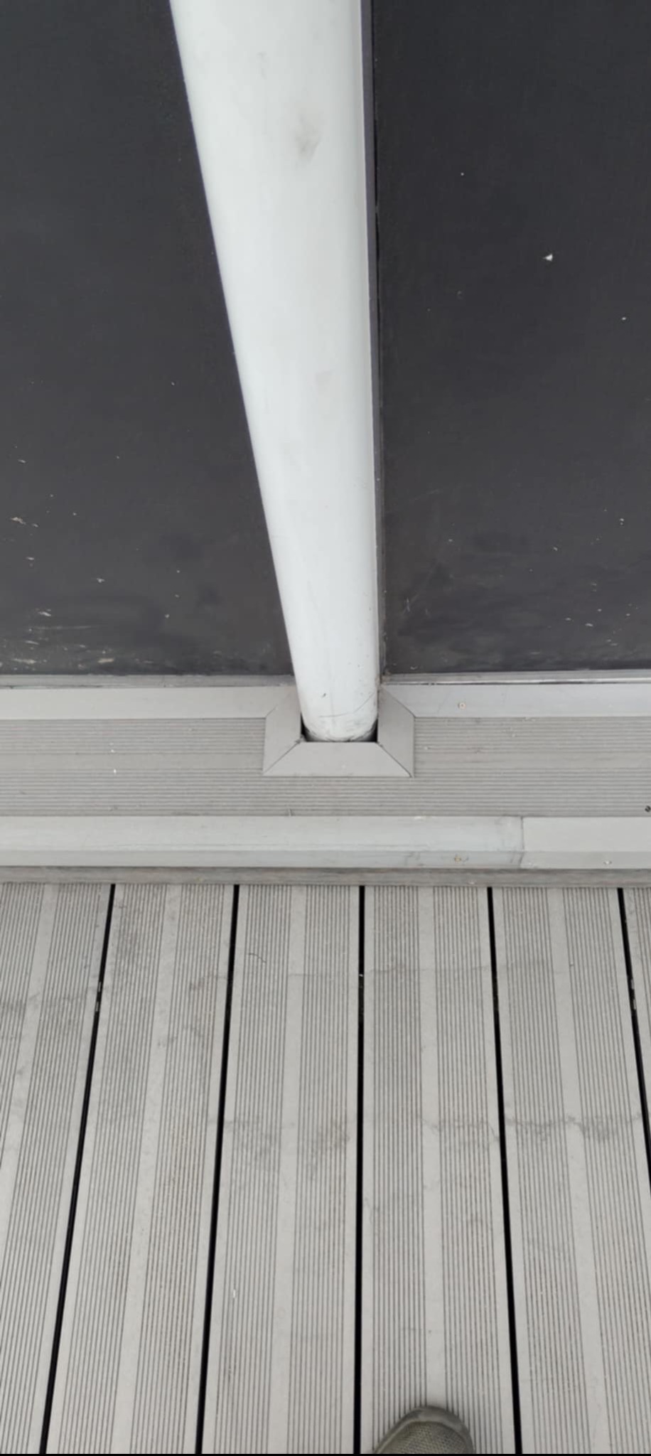 Szare deski tarasowe kompozytowe z czarnymi wstawkami, biała pionowa rura pod czarnym sufitem, widok z dołu, perspektywa z lekkim skosem.