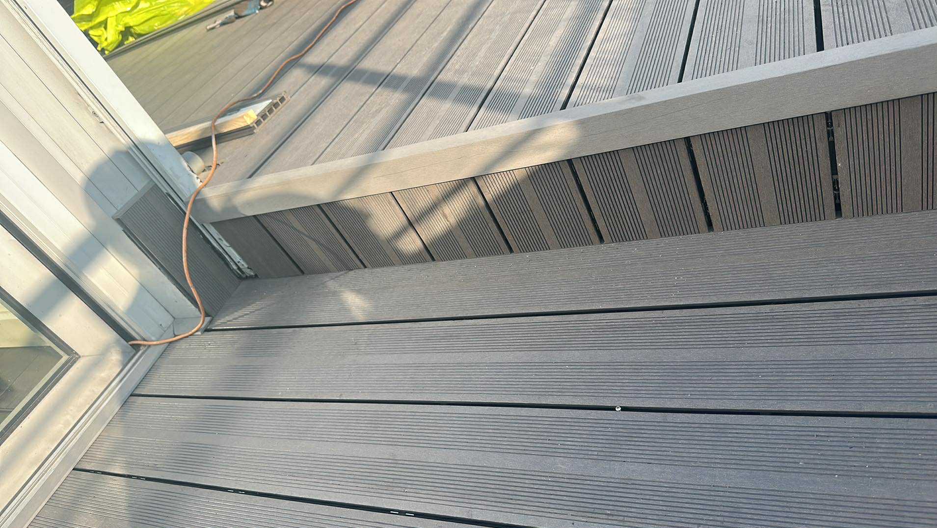 Szare deski tarasowe kompozytowe ułożone na balkonie, widoczne z bliska. Deski z wyraźnym wzorem, przy oknie balkonowym. Kabel elektryczny biegnie wzdłuż ramy okiennej.