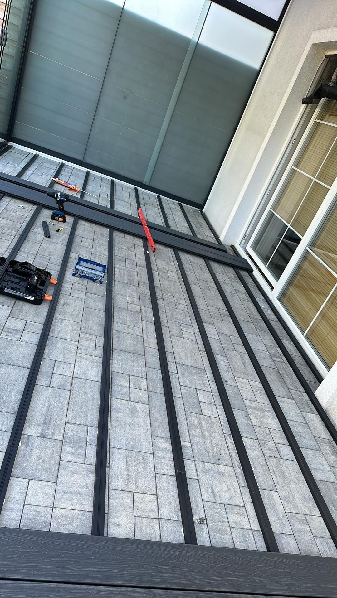 Montaż konstrukcji tarasu w Warszawie: Widok z góry na szare płyty i czarne profile, narzędzia pracy widoczne w kadrze, okno w tle.