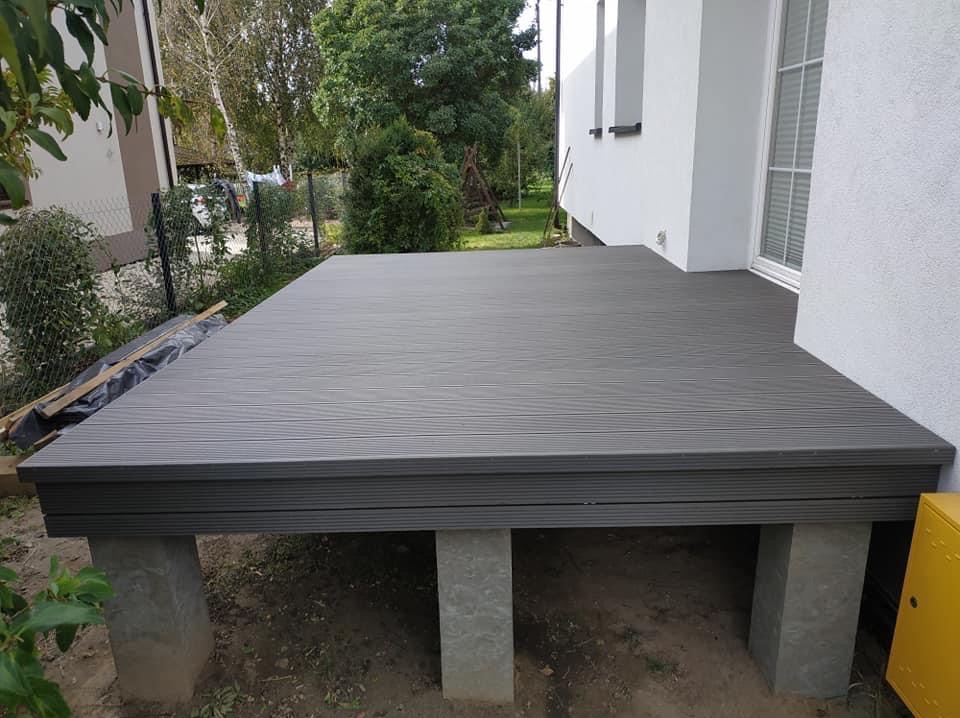 Szary taras na betonowych podporach przy białym budynku. Deski z wyraźnym wzorem drewna. Widoczne elementy ogrodu i żółta skrzynka gazowa.