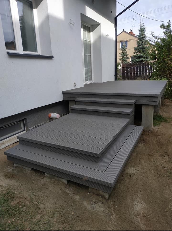 Szary taras kompozytowy z schodami przy białym domu, widok z boku. Widoczne deski tarasowe i betonowe podpory. Dom z oknami w tle, pochmurne niebo.