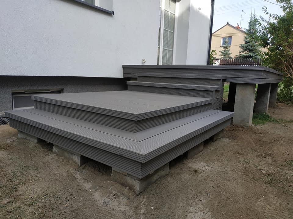 Nowoczesny taras kompozytowy w odcieniach szarości, z wyraźną strukturą desek, połączony ze schodami i fragmentem podwyższonej platformy na betonowych podporach.