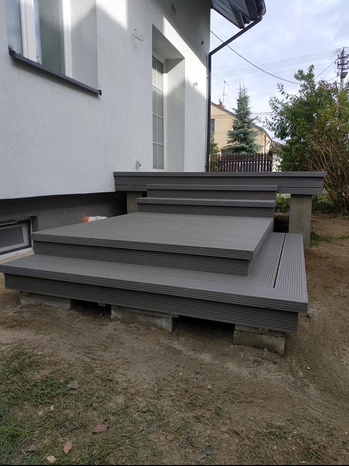 Schody wejściowe z szarej deski kompozytowej, prowadzące do białego domu. Widoczne betonowe podpory i ziemiste otoczenie. Ujęcie z dołu.