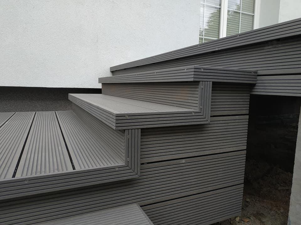 Schody z desek kompozytowych w odcieniach szarości, prowadzące do domu z białymi ścianami. Widoczne detale montażu i faktura materiału.