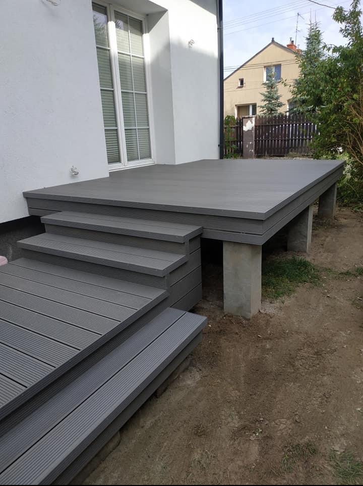 Szary taras kompozytowy z schodami, wsparty na betonowych filarach, przylegający do białego budynku z oknem. Widok z boku, ziemia w tle.