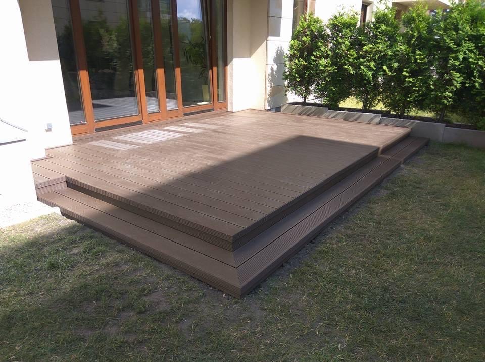 Nowoczesny taras kompozytowy z dwoma stopniami, brązowe deski, widok z poziomu trawnika, budynek z dużymi oknami w tle, krzewy.