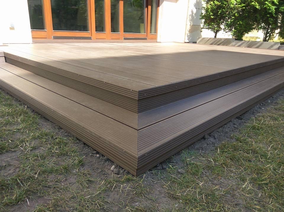 Nowoczesny taras kompozytowy z brązowych desek, z widocznymi schodkami, na tle trawnika i budynku z dużymi oknami, prezentujący solidne wykonanie.