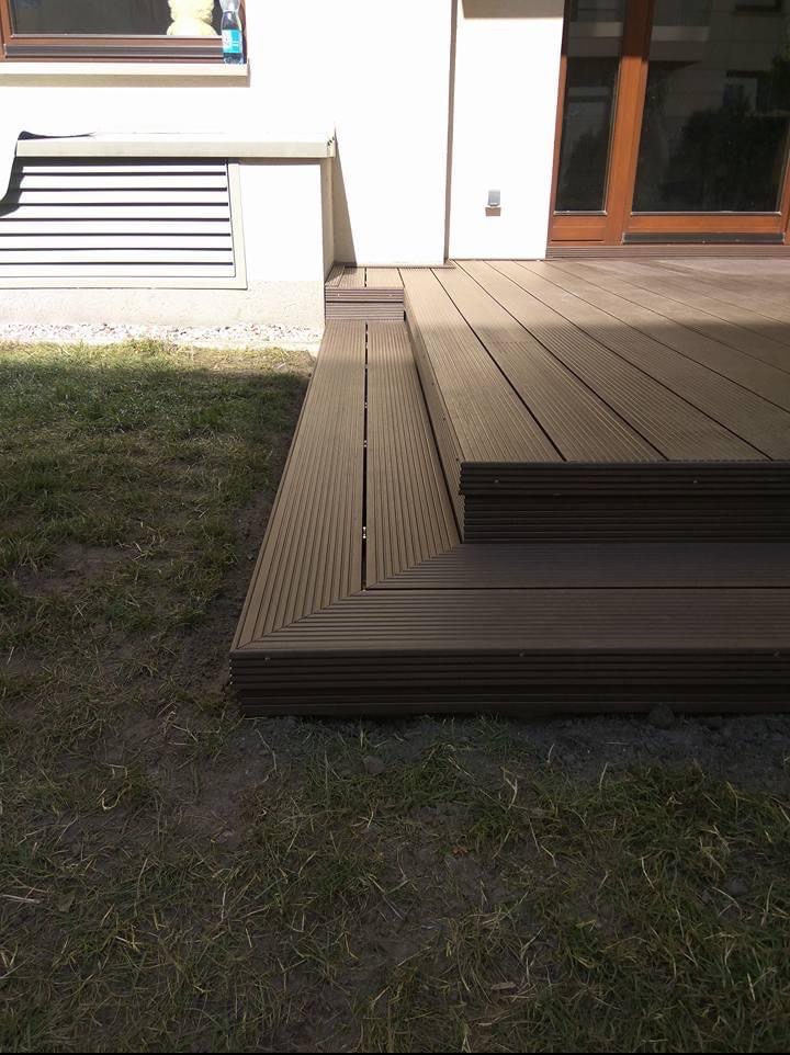 Nowoczesny taras z desek kompozytowych w odcieniach brązu, zintegrowany ze schodami wejściowymi do budynku. Widok na trawnik i fragment elewacji.