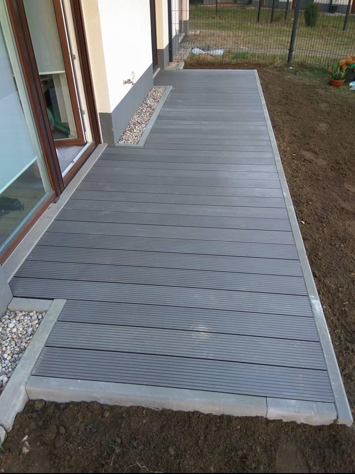 Szary taras kompozytowy z obrzeżem z kostki betonowej, przylegający do budynku z oknem. Widoczne drobne kamienie ozdobne i ziemia.