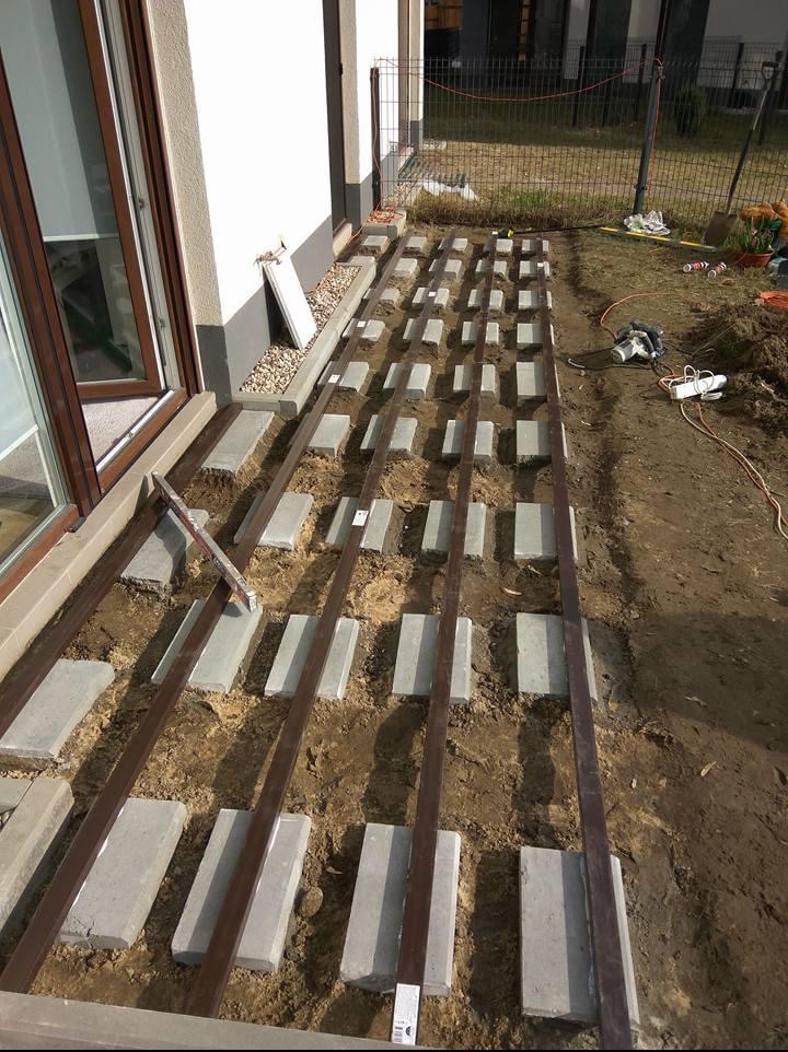 Przygotowanie podłoża pod taras: betonowe bloczki ustawione w rzędach, metalowe legary, ziemia. Widoczne narzędzia i fragment budynku.