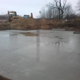 Wylana płyta fundamentowa z wystającymi prętami zbrojeniowymi. W tle koparki i wykop pod fundamenty. Pochmurny dzień, teren budowy po deszczu.