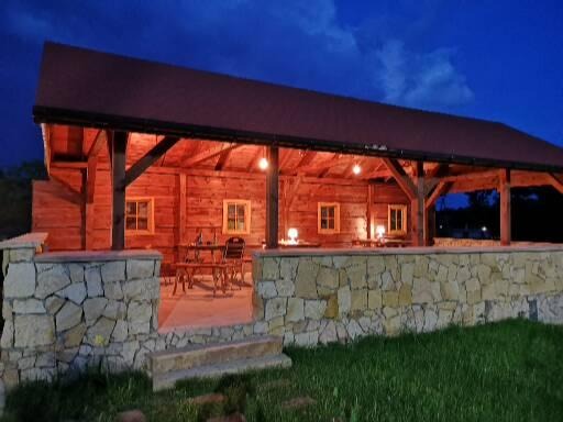 Drewniana altana z kamienną podmurówką oświetlona wieczornym światłem, z widocznym stołem i krzesłami wewnątrz.