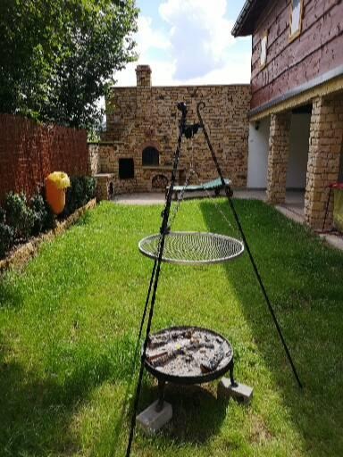 Ogród z murowanym grillem i paleniskiem na trójnogu, z resztkami popiołu, w tle kamienny piec chlebowy i budynek z drewnianą elewacją.