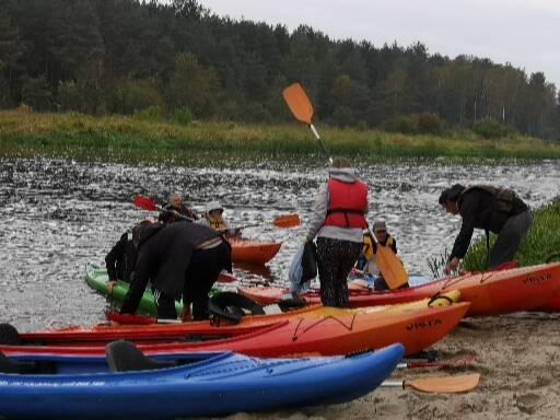 Grupa osób przygotowuje się do spływu kajakowego na rzece, widoczne kolorowe kajaki na brzegu i uczestnicy z wiosłami, ubrani w kamizelki ratunkowe.