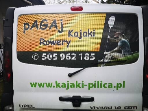 Tył białego Opla Vivaro z reklamą wypożyczalni kajaków: grafika z osobą płynącą kajakiem, napisy 'PAGAJ Kajaki Rowery', numer telefonu i adres strony internetowej 'www.kajaki-pilica.pl'.