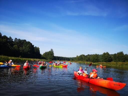 Grupa osób płynąca kajakami po rzece podczas imprezy integracyjnej, widoczne lasy na brzegach i błękitne niebo.