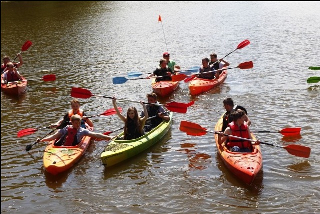 Grupa osób płynie kajakami po rzece podczas słonecznego dnia. Widoczne kolorowe kajaki i wiosła. Jeden z kajaków jest zielony, pozostałe pomarańczowe. W tle drzewa i spokojna woda.