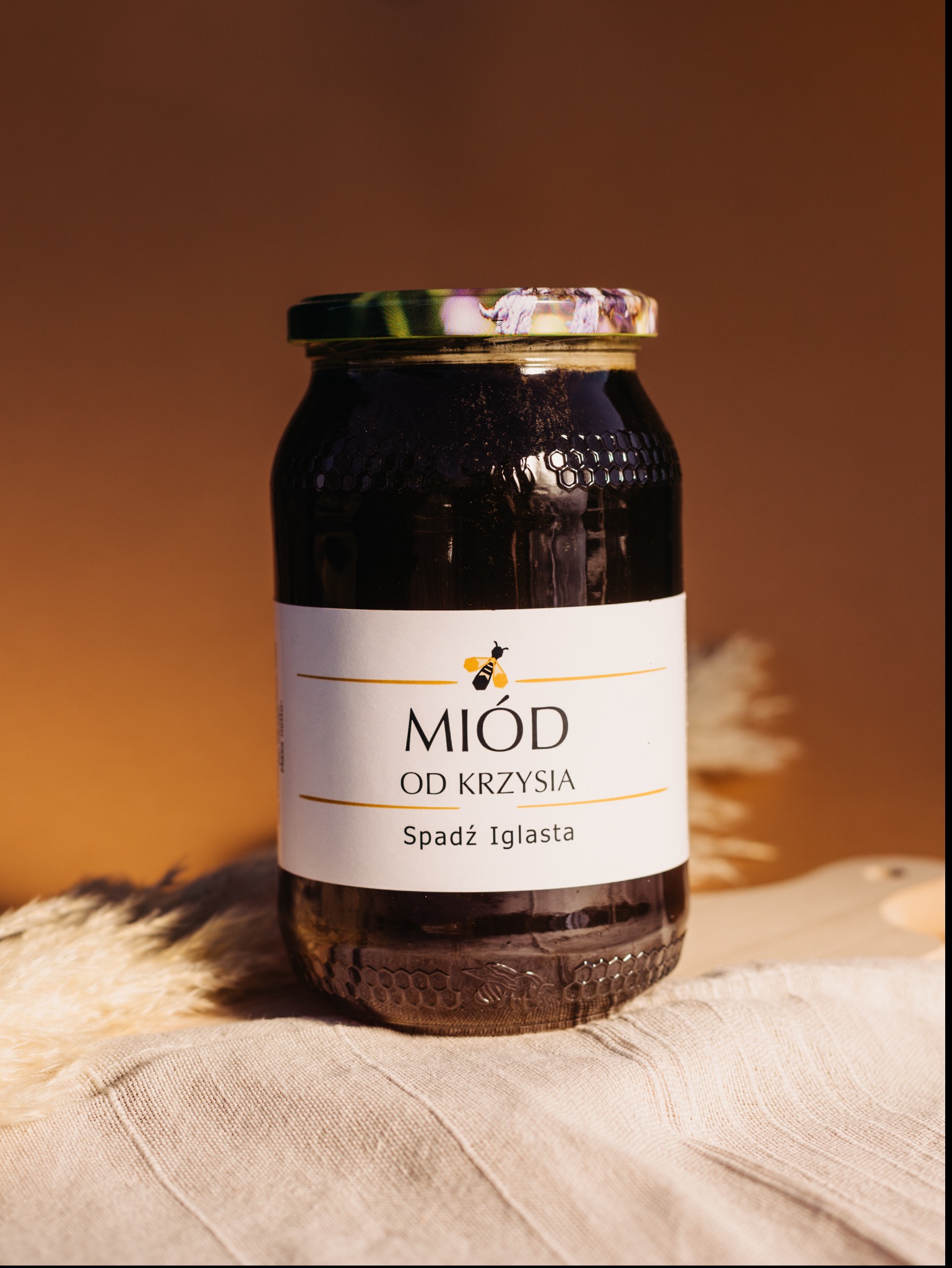 Słoik ciemnego miodu spadziowego z etykietą 'Miód od Krzysia', umieszczony na lnianej ściereczce i drewnianej desce, z brązowym tłem.