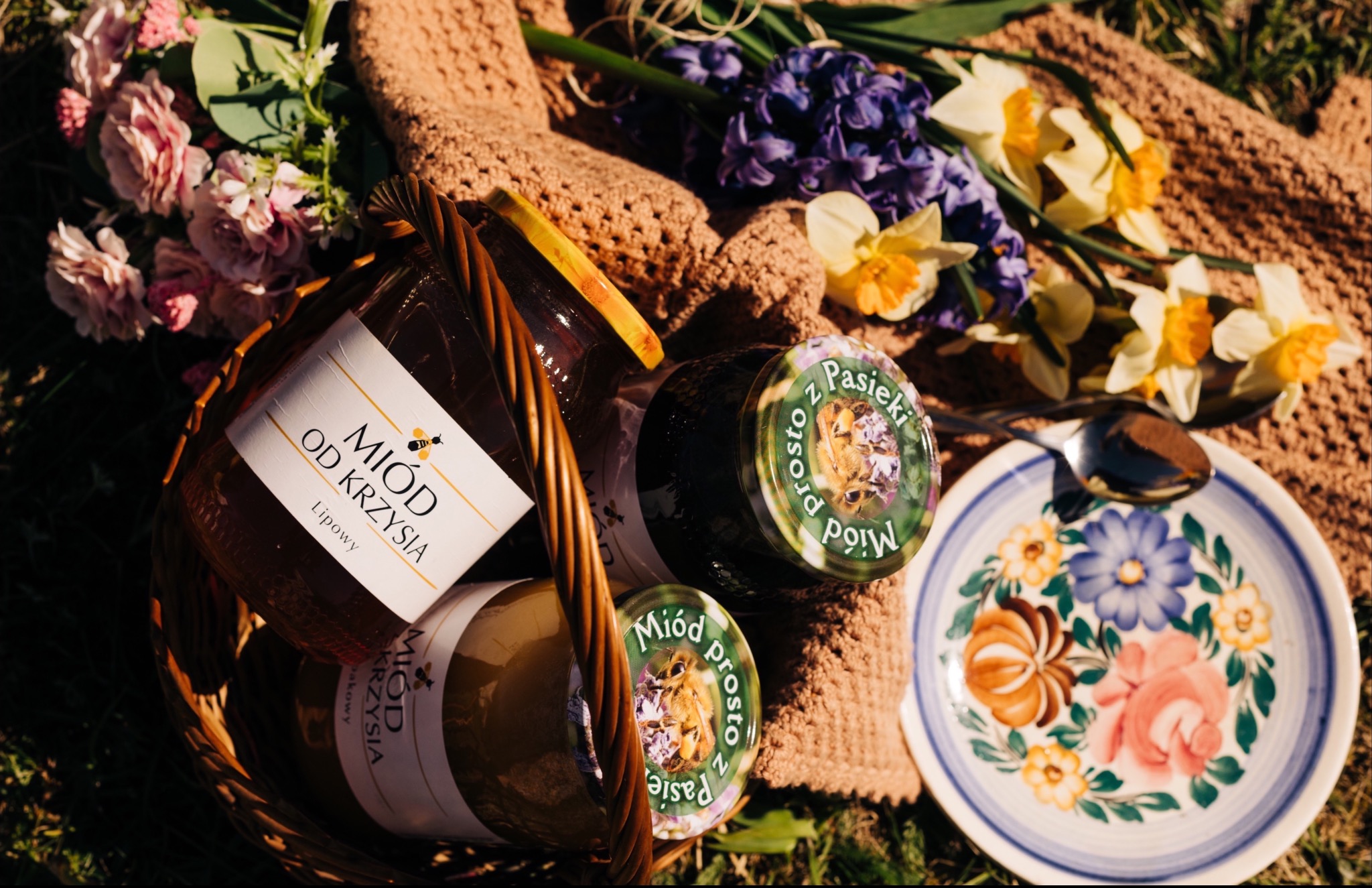Kompozycja z wiklinowego kosza wypełnionego słoikami z miodem od różnych producentów, otoczona wiosennymi kwiatami i talerzem z łyżeczką, ułożona na brązowej dzierganej serwecie na trawie.