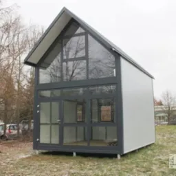 Nowoczesny, mały dom z dużymi przeszkleniami w szarej ramie, na tle drzew bez liści i pochmurnego nieba, widoczny z boku, z logo firmy w prawym dolnym rogu.