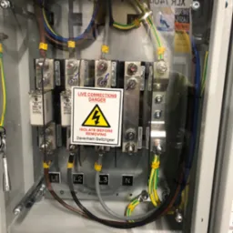 Otwarta skrzynka elektryczna z podłączonymi przewodami oznaczonymi L1, L2, L3 i N, widoczne bezpieczniki Bussmann i ostrzeżenie o napięciu 'Live Connections Danger' Davenham Switchgear.