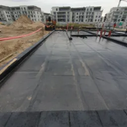 Czarna, płaska powierzchnia dachu pokryta papą termozgrzewalną w trakcie prac, widoczne kominki wentylacyjne, w tle budynki mieszkalne i koparka.