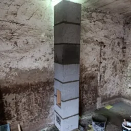 Wznoszona konstrukcja komina z szarych bloczków betonowych, z widocznymi spoinami zaprawy, w surowym wnętrzu budynku w trakcie budowy, z widocznym otworem w stropie i narzędziami murarskimi obok.