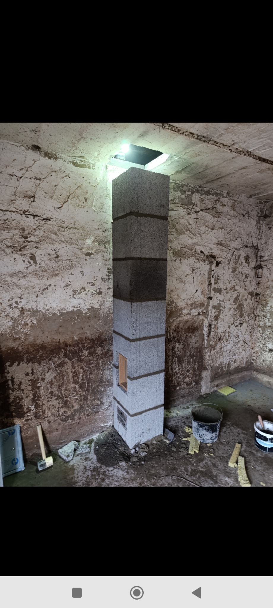 Wznoszona konstrukcja komina z szarych bloczków betonowych, z widocznymi spoinami zaprawy, w surowym wnętrzu budynku w trakcie budowy, z widocznym otworem w stropie i narzędziami murarskimi obok.