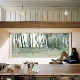 Wnętrze pokoju z drewnianym stołem i oknem wnękowym, w którym siedzi mężczyzna, widok na las, minimalistyczne oświetlenie.