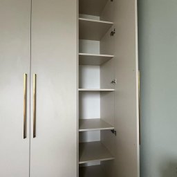 Fabis Meble na Wymiar - Wysoka, beżowa szafa z otwartymi drzwiami ukazującymi puste półki, złote, pionowe uchwyty na zamkniętych drzwiach po lewej stronie, widoczne zawiasy.