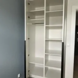 Wnętrze białej, otwartej szafy wnękowej z półkami i drążkiem na ubrania, widoczne zawiasy i fragment ściany w kolorze gołębim.