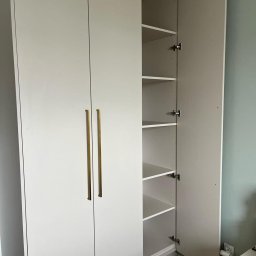 Fabis Meble na Wymiar - Biała szafa wnękowa z otwartymi drzwiami ukazującymi półki, z długimi złotymi uchwytami, na tle zielonej ściany i parkietu.
