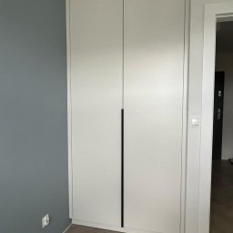 Fabis Meble na Wymiar - Biała, dwudrzwiowa szafa z minimalistycznymi, długimi, czarnymi uchwytami, wkomponowana w narożnik pokoju z szarą ścianą i parkietem ułożonym we wzór jodełki.