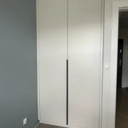 Biała, dwudrzwiowa szafa z minimalistycznymi, długimi, czarnymi uchwytami, wkomponowana w narożnik pokoju z szarą ścianą i parkietem ułożonym we wz&oacute;r jodełki.