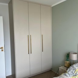 Fabis Meble na Wymiar - Trzydrzwiowa, beżowa szafa na wymiar z długimi, złotymi uchwytami, stojąca w sypialni z zieloną ścianą i parkietem ułożonym w jodełkę.