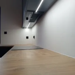 Fragment blatu kuchennego z płyty drewnopodobnej, płyta indukcyjna, oświetlenie LED podszafkowe i czarne gniazdka elektryczne na białej ścianie.