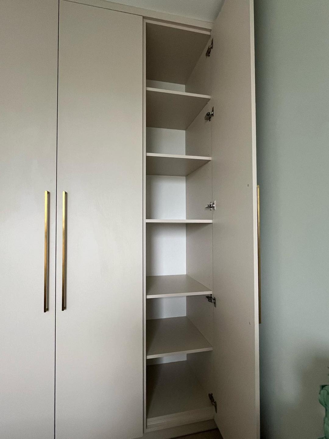 Wysoka, beżowa szafa z otwartymi drzwiami ukazującymi puste półki, złote, pionowe uchwyty na zamkniętych drzwiach po lewej stronie, widoczne zawiasy.