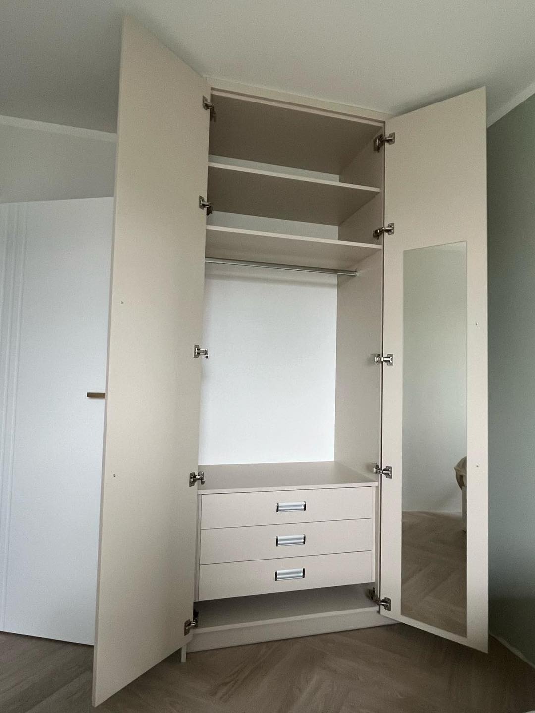 Otwarta, beżowa szafa z trzema szufladami, półkami i drążkiem na wieszaki; jedna z drzwi wyposażona w lustro.