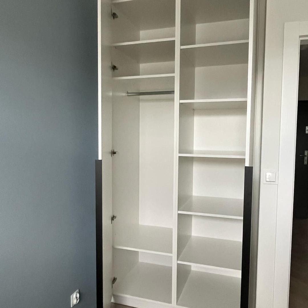 Wnętrze białej, otwartej szafy wnękowej z półkami i drążkiem na ubrania, widoczne zawiasy i fragment ściany w kolorze gołębim.