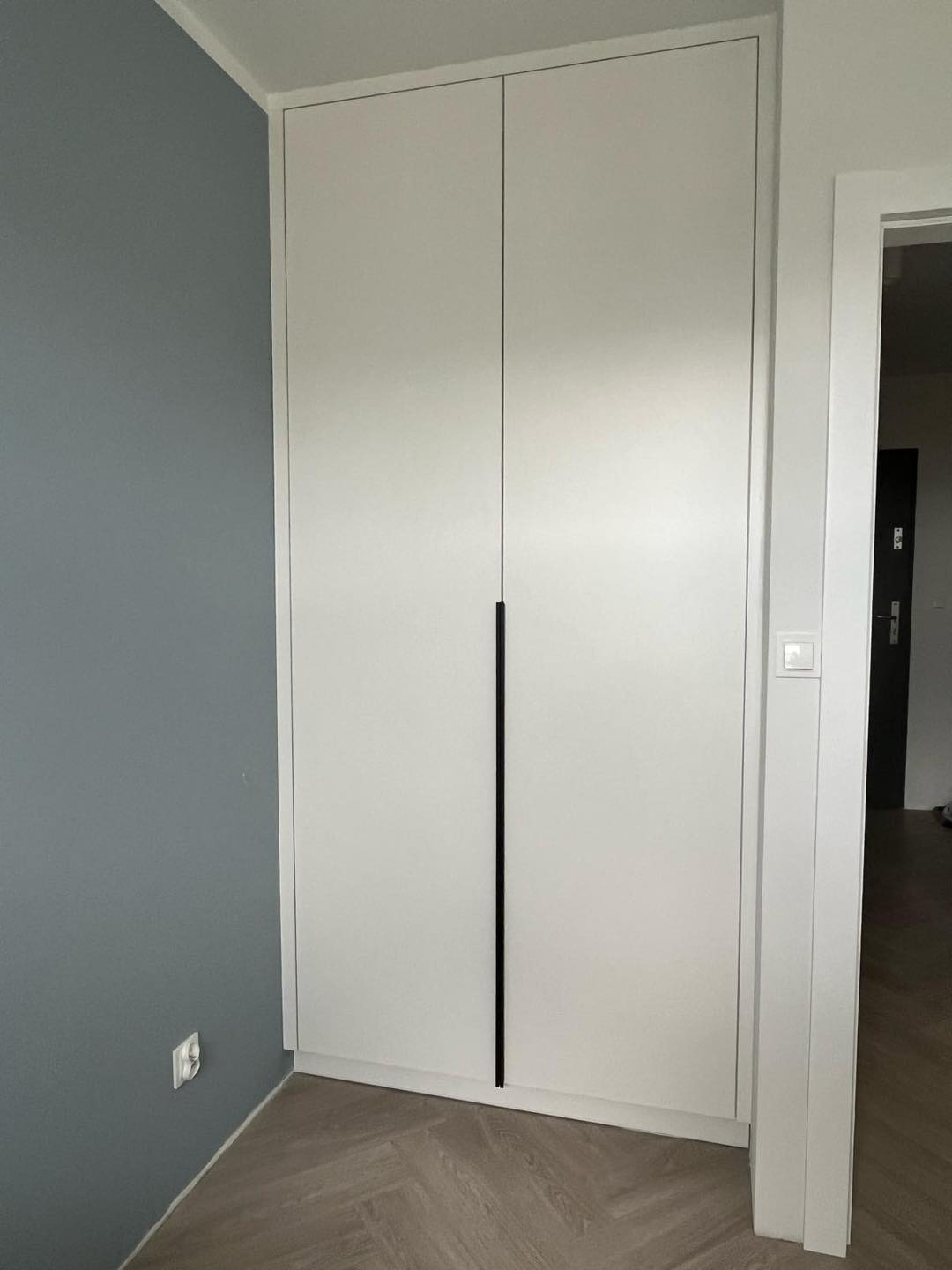 Biała, dwudrzwiowa szafa z minimalistycznymi, długimi, czarnymi uchwytami, wkomponowana w narożnik pokoju z szarą ścianą i parkietem ułożonym we wzór jodełki.