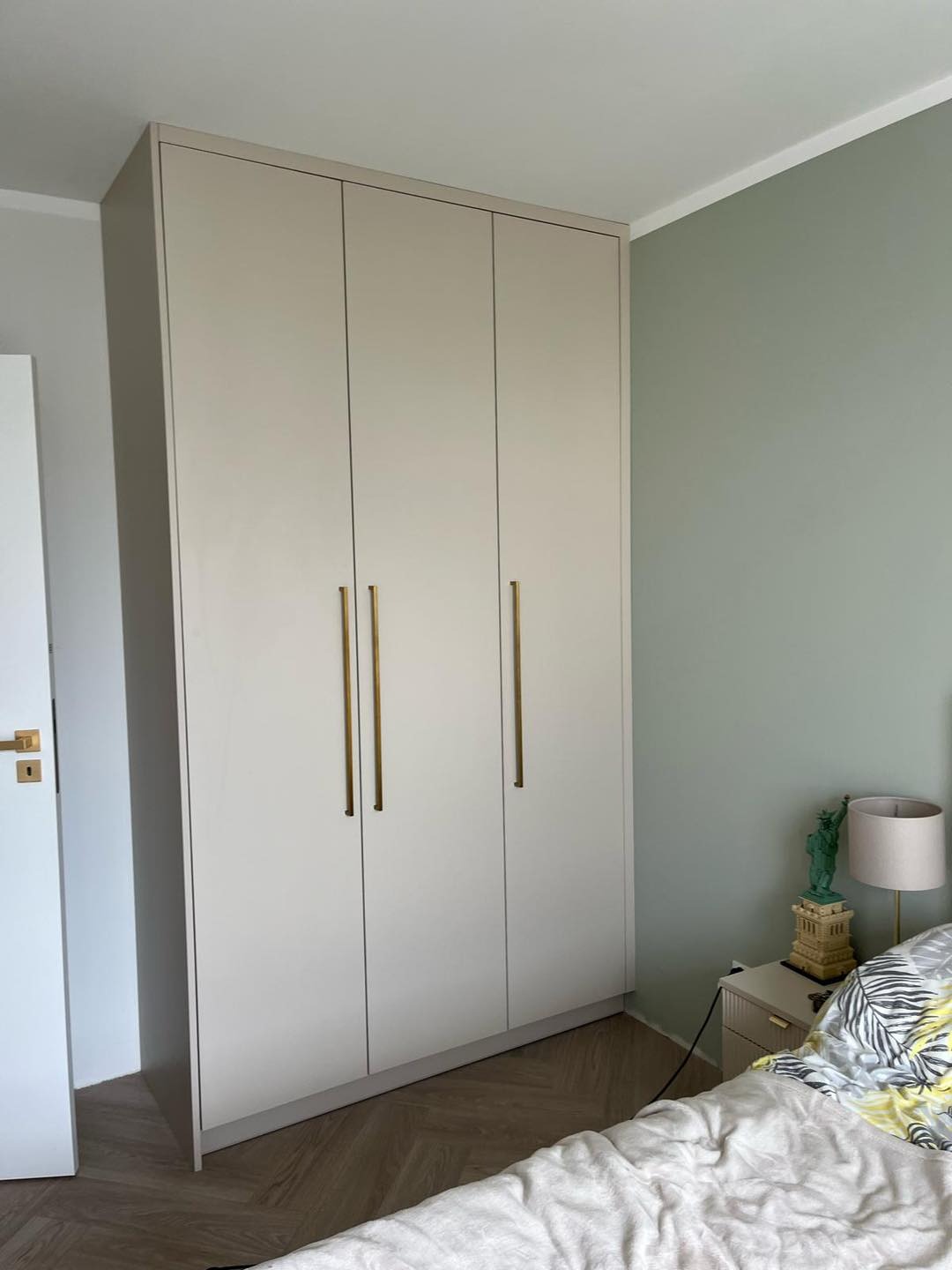 Trzydrzwiowa, beżowa szafa na wymiar z długimi, złotymi uchwytami, stojąca w sypialni z zieloną ścianą i parkietem ułożonym w jodełkę.