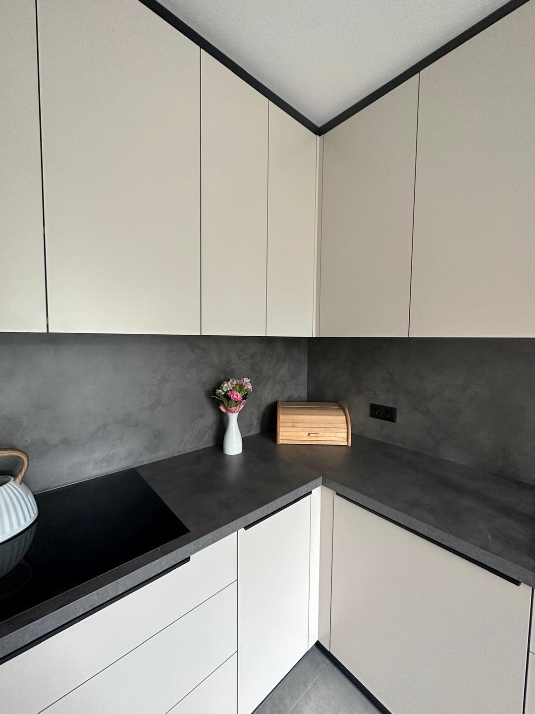 Nowoczesna kuchnia w odcieniach bieli i szarości z minimalistycznymi frontami, ciemnym blatem i drewnianym chlebakiem jako akcentem dekoracyjnym.