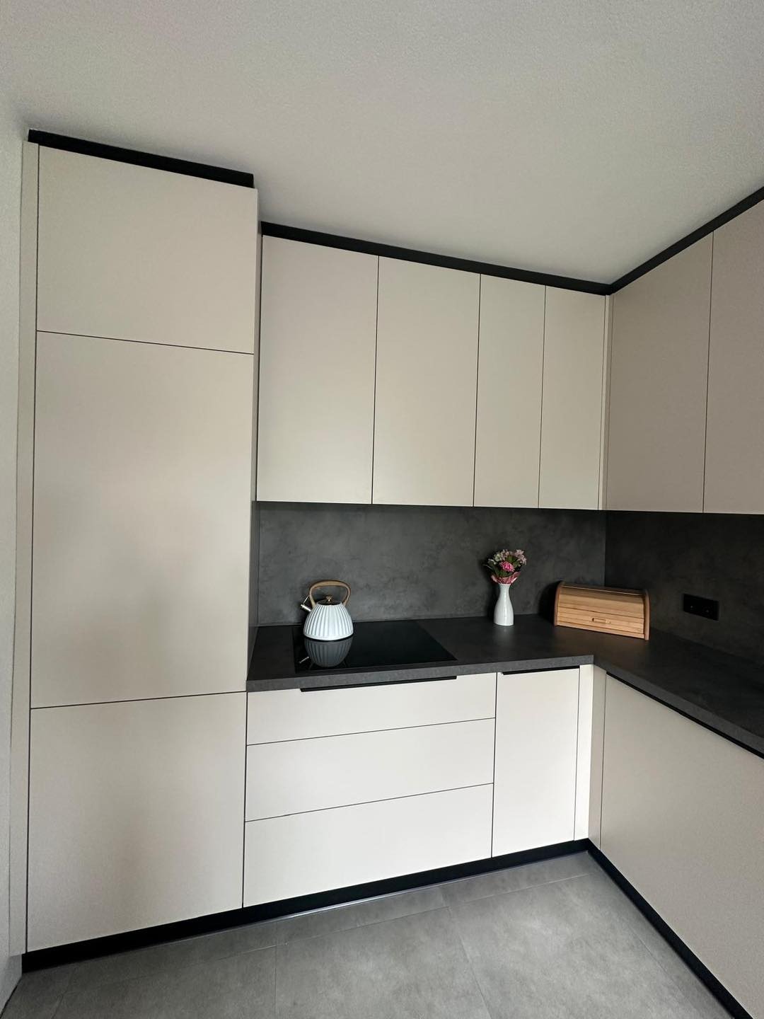 Nowoczesna kuchnia z jasnymi, gładkimi frontami szafek, czarnymi akcentami i szarym blatem. Na blacie czajnik w stylu retro i wazonik z kwiatami.