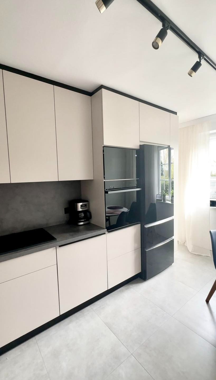 Nowoczesna kuchnia w beżowych odcieniach z czarnymi akcentami, oświetleniem punktowym na szynie i sprzętem AGD w zabudowie, widok na okno z firanką.