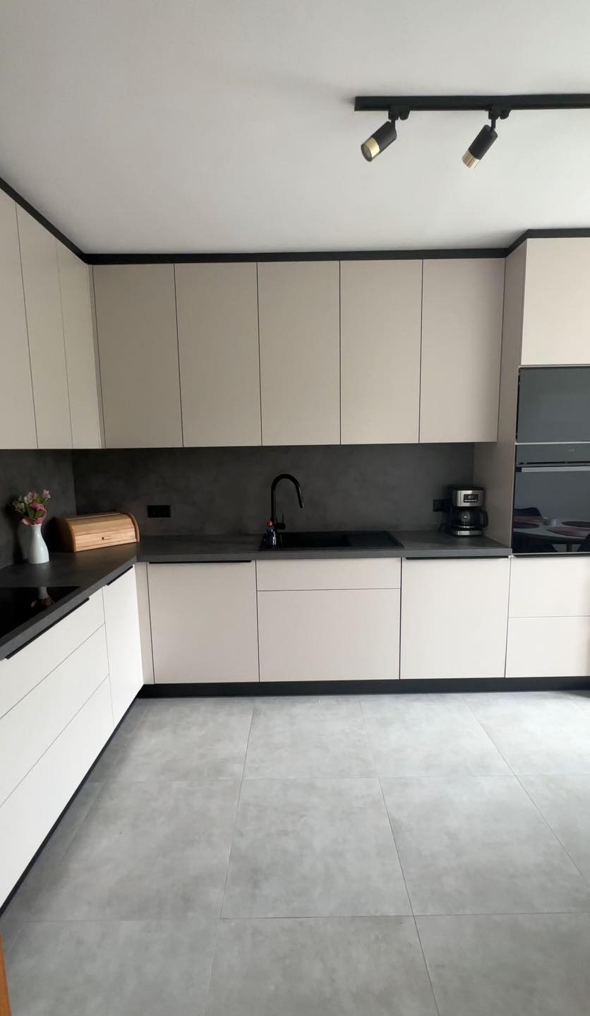 Jasna kuchnia z ciemnymi akcentami: czarne obramowania szafek, bateria i oświetlenie punktowe. Na blacie drewniany chlebak i wazon z kwiatami.