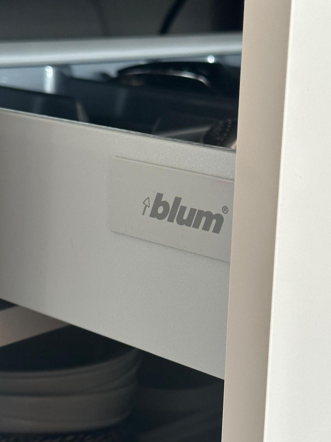 Szary napis 'blum' na białej szufladzie z widoczną zawartością, wewnątrz szafki.