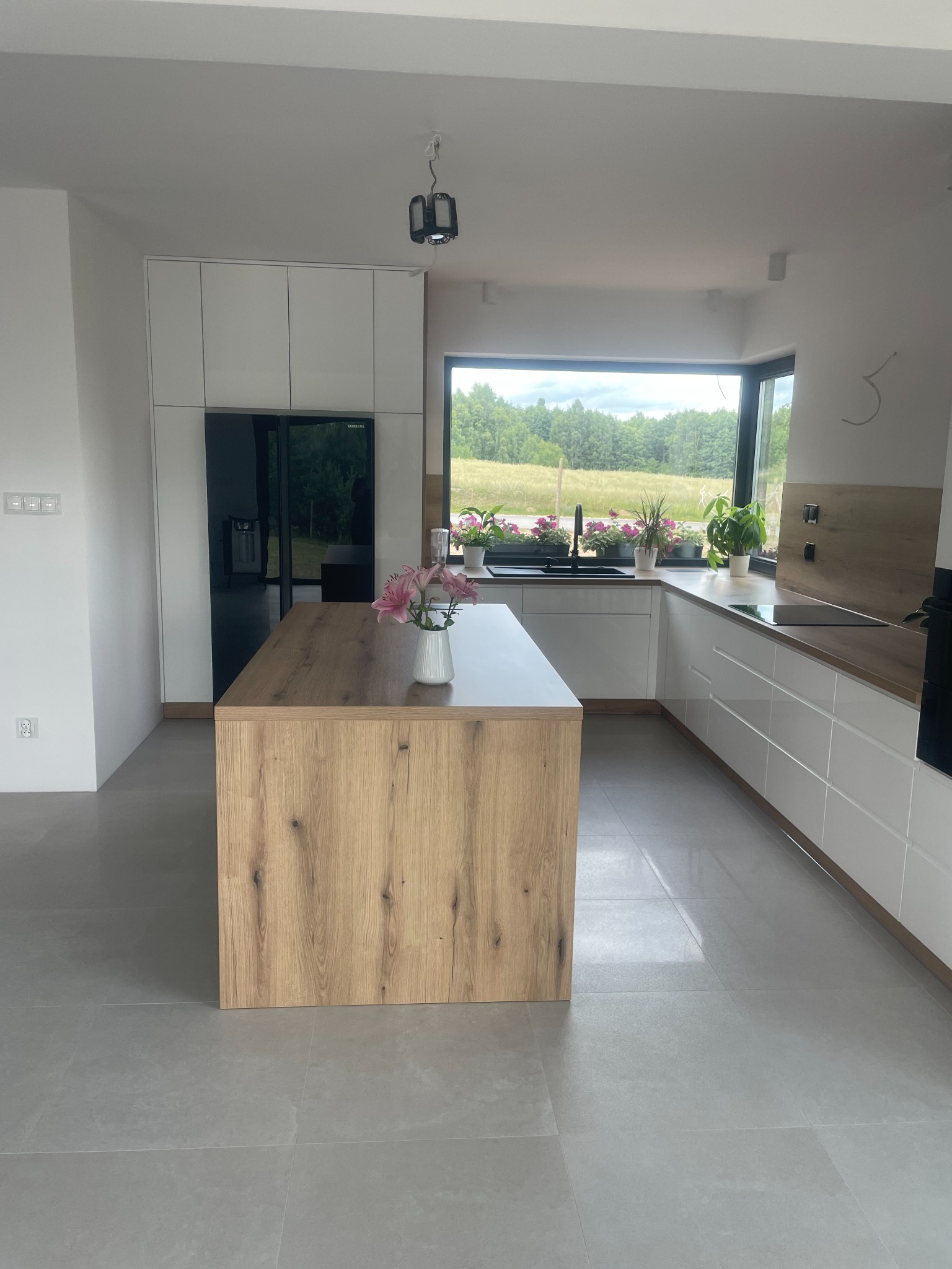 Nowoczesna kuchnia z wyspą o drewnianym blacie i białymi szafkami, widok na zielony ogród przez duże okno.