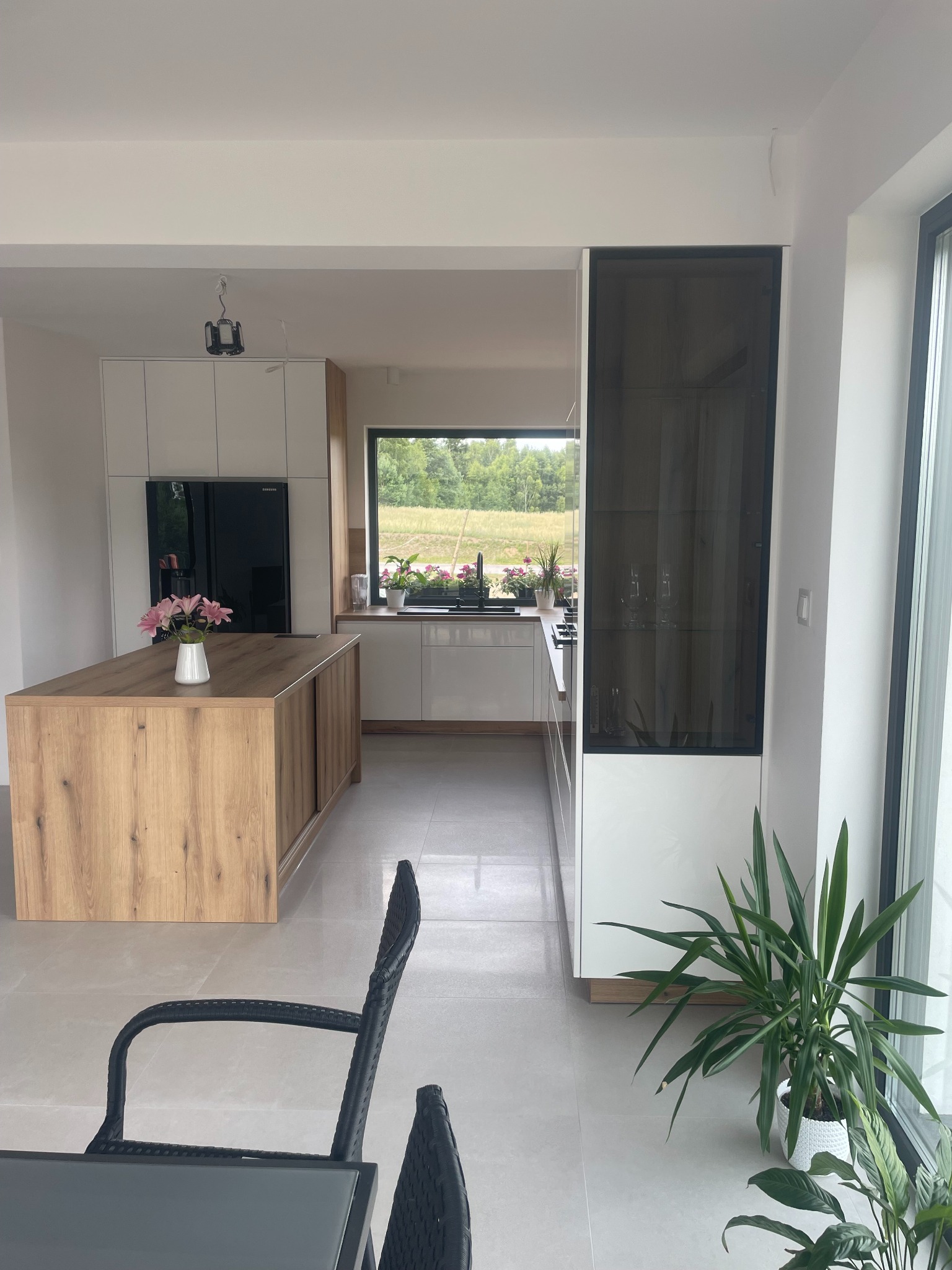 Nowoczesna kuchnia z wyspą o drewnianym wykończeniu, białymi szafkami i czarnym sprzętem AGD, widok z jadalni, z kwiatami na wyspie i rośliną doniczkową przy oknie.