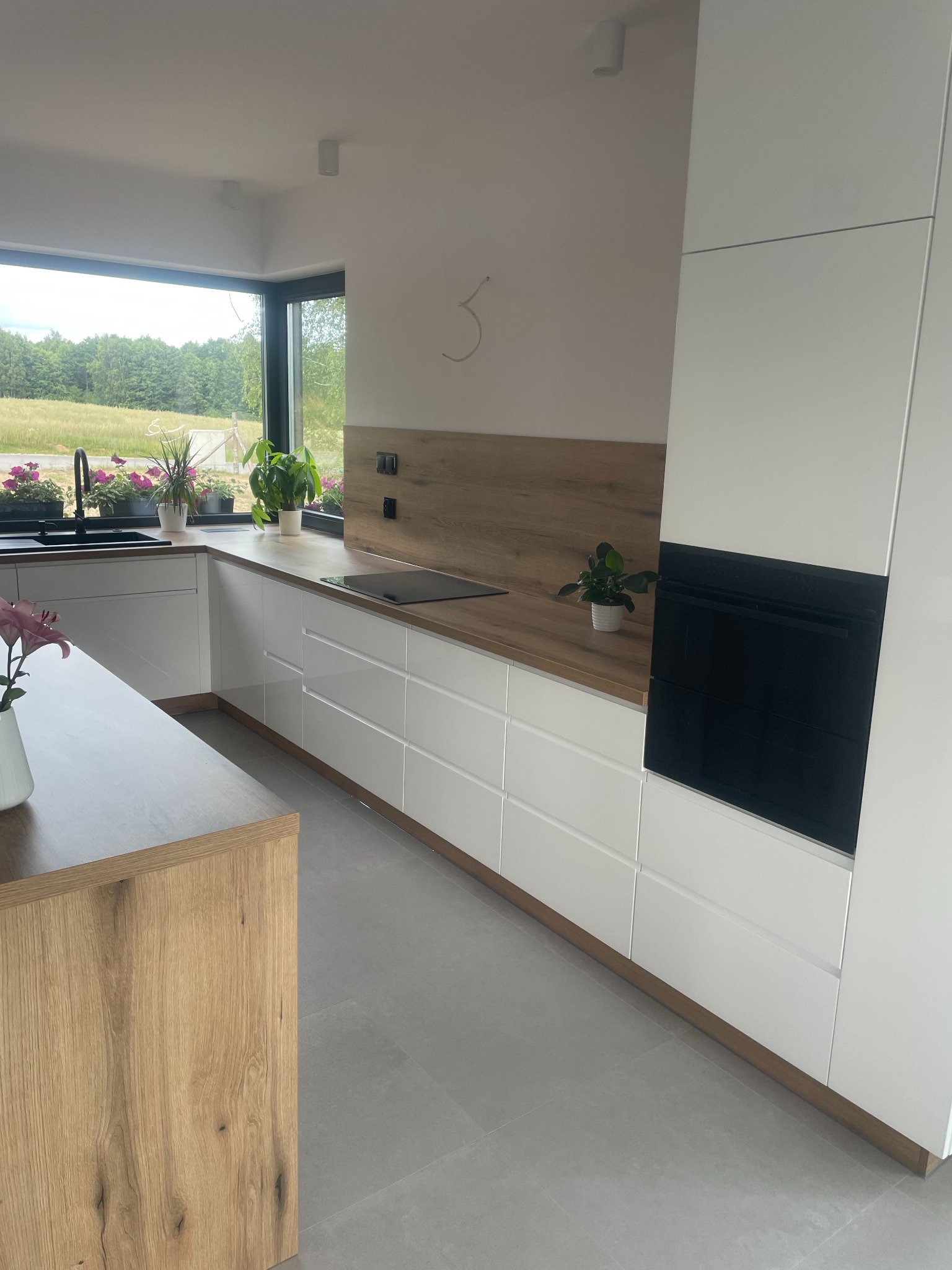 Nowoczesna, jasna kuchnia z białymi frontami i drewnianym blatem, z widokiem na ogród przez duże okno. Minimalistyczny design z czarnym piekarnikiem w zabudowie i roślinami doniczkowymi na blacie.